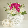 A chic wedding-ready floral design is captured in gentle, diffused light, as if placed near a bright window in a Queensbury home. At the centre sits a lush knot of fuchsia blooms-full-petalled peonies or roses with deep, velvety texture-drawing the gaze like a jewel. Around this glowing core, creamy ivory alstroemeria open in soft stars, their trumpet-shaped petals delicately marked with fine magenta streaks and touches of green, adding a sense of artistry and fine detail. The flowers are nestled tightly into a perfectly round white wicker container, whose smooth, interlaced bands form a dazzling, basket-like bowl. Its geometric pattern feels clean and contemporary, while the handwoven texture whispers of tradition, much like the mix of old and new you see walking between Queensbury Station and the nearby residential streets. Fresh green leaves peek between the blooms, some gently arched, others upright and youthful, suggesting new beginnings and the unfolding of shared stories. Two cream-coloured wicker orbs rest beside the arrangement, their airy, spherical shape mirroring the basket and lending a sculptural calm to the composition. The overall impression is one of lightness and intention: a piece designed to sit gracefully on a wedding signing table, an intimate reception buffet, or a quiet mantelpiece at home after the celebration. You can almost sense the soft rustle of petals and the cool, green scent when the stems are first trimmed. Created with the care of an experienced local florist, this arrangement suits brides and couples in Queensbury seeking understated elegance and romance, with easy local delivery for those meaningful moments.
