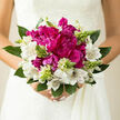 In this photograph, a romantic bridal bouquet is captured in calm, natural daylight, its colours and textures echoing the relaxed elegance of a Queensbury wedding. At the centre, full magenta peonies sit like little clouds of satin, their many layers of petals soft, frilled, and almost weightless, as if a breeze from nearby Kingsbury Road might stir them at any second. They bring a bold yet tender warmth to the arrangement, suggesting passion held with gentleness. Surrounding them, white alstroemeria blooms form a graceful halo, each petal edged with faint ripples and traced with fine burgundy lines that look hand-painted. These markings invite the eye to linger, softening the intensity of the peonies and adding a sense of intricate detail. Threaded through the bouquet are slender stems tipped with pale green buds, catching the light and lending a sense of new beginnings, like fresh shoots emerging after a spring rain in one of Queensbury's quiet residential gardens. Layered greenery supports the flowers, the leaves broad and cool, curving around the base to cradle the whole design. Their deep green tones anchor the bouquet and highlight the brightness of the magenta and white blooms. The overall mood is serene yet quietly joyful, a bouquet meant for a bride stepping out of a classic car in Queensbury, ready to walk into a church or registry office with calm anticipation. You can almost sense the subtle scent-a mix of soft floral sweetness and leafy freshness-drifting gently in the air as this carefully crafted bouquet rests in waiting hands, prepared by local florists for same-day wedding delivery.