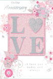 An anniversary card is displayed in close view, revealing an intricate design that balances romance and refinement with a softness that feels perfectly suited to a quiet celebration in Queensbury. At its heart, the word LOVE is prominently featured, rendered in raised, blush-pink letters edged in delicate, shimmering glitter. The slightly embossed texture makes the word feel almost sculpted, akin to the way a florist might carefully position the focal flowers in a bouquet to give them extra presence. Surrounding this central word, a ring of illustrated blossoms and foliage spreads outward, drawn in fine, flowing lines. The flowers appear airy and stylised, shaded with blush, ivory, and pale silver, their petals curling gently as if stirred by a light breeze over a garden off Kenton Road. Slender vines and leaves weave between the blooms, some kissed with silver foil that catches the light like tiny dewdrops. The background of the card is a soft, powdery pink, just a touch deeper behind the central motif and fading to a paler tone at the edges, creating a soothing, dreamlike effect. Tucked above and below the floral arrangement is an elegant script message, offering loving anniversary sentiments in understated grey ink that harmonises with the card's gentle palette. The overall composition feels as thoughtful as an anniversary bouquet hand-delivered around Queensbury-subtle, heartfelt, and designed to be treasured as a reminder of shared years and the promise of many more to come.